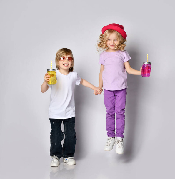Little girl and boy in colorful casual clothes. Smiling, holding hands, yellow and pink cocktail bottles. Posing isolated on white - Photo, Image