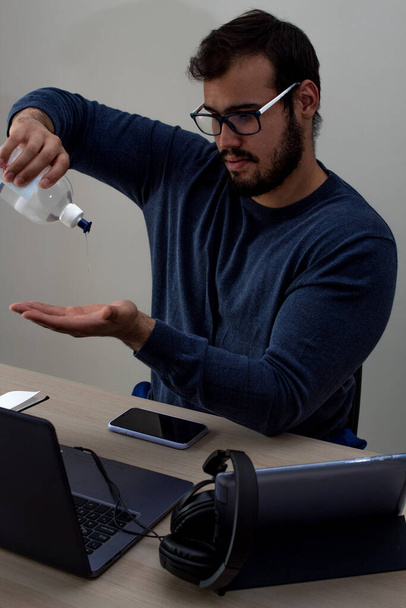 Young caucasian man with glasses and a blue sweater applying hand sanitizer before starting his workday with a laptop and his tablet with a phone with headphones - Photo, image