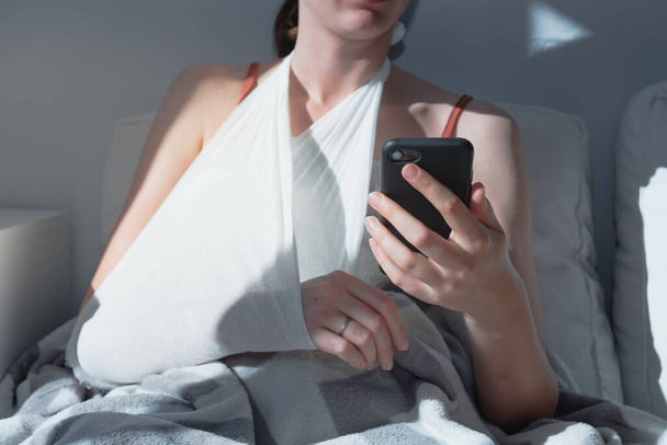 Wounded arm in bondage, woman rests in  bed, body parts. Caucasian female adult in bright sun-lit room holds a phone, concept of self treatment, medical treatment at home - Фото, изображение