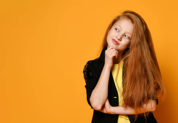 Ginger teenage child in black jacket and pants, yellow t-shirt. She is looking thoughtful, posing on orange background. Close up - Valokuva, kuva