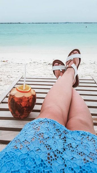 Eine junge Frau entspannt sich und sonnt sich auf einer Liege in Strandnähe und trinkt einen Kokoscocktail. Blaues Spitzenkleid, weißer Sand und kristallklares Meerwasser. Luxusurlaub auf tropischen Inseln. - Foto, Bild