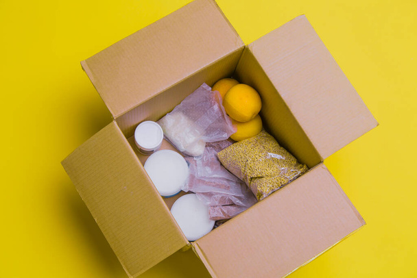 the main products for self-isolation in a box: cereals, buckwheat, fruit, canned food on a yellow background. home delivery. Assistance to population - Fotografie, Obrázek