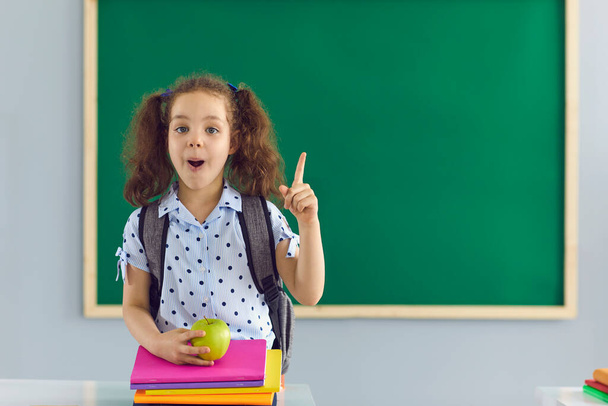 Cute girl with pile of books and apple pointing up near chalkboard at classroom. Little child having idea at school - Photo, image