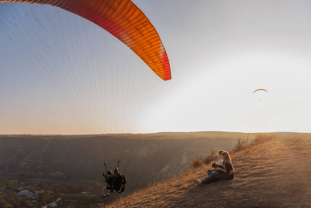 Happy couple in love are resting on the mountainside with a black Schnauzer, sitting on the ground, hugging a dog, watching bright  paragliders fly in the blue sky, admire the sunset, travel with a dog - Zdjęcie, obraz
