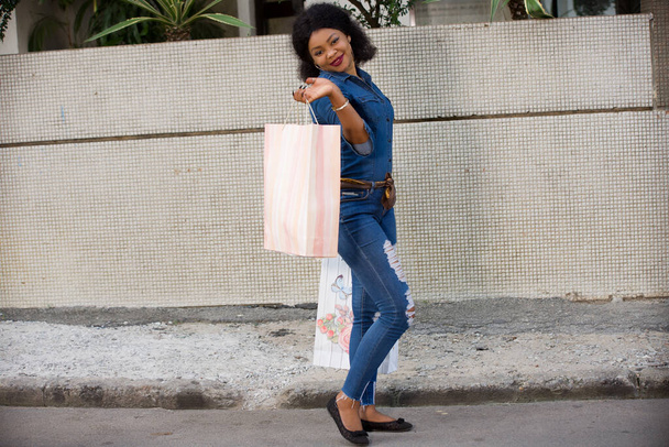 Ελκυστική νεαρή ευτυχισμένη Αφροαμερικανή γυναίκα περπατώντας σε ένα αστικό περιβάλλον της πόλης και μεταφέροντας τσάντες για ψώνια. - Φωτογραφία, εικόνα