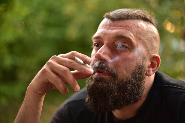 Man with a beard on a background of greenery. Large facial portrait of a bearded man of 40 years. Portrait full face. Man in a black T-shirt. A man smokes a cigarette. - Photo, Image