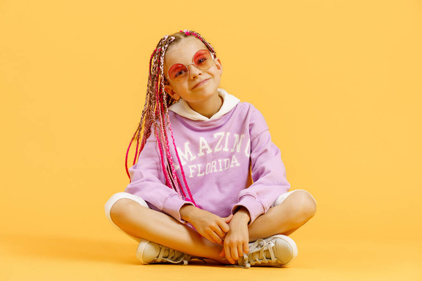 Stylish girl in rounded glasses with pink dreadlocks sitting, looking at camera and posing on a yellow background. Beauty, fashion - Photo, image