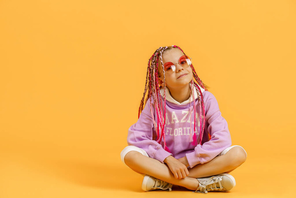 Stylish girl in rounded glasses with pink dreadlocks sitting, looking at camera and posing on a yellow background. Beauty, fashion - Photo, image