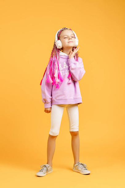 Adorable child in rounded glasses with pink dreadlocks listen to music playing in earphones and dance on yellow background. Cute small kid listening to music in headset, copy space - Fotó, kép