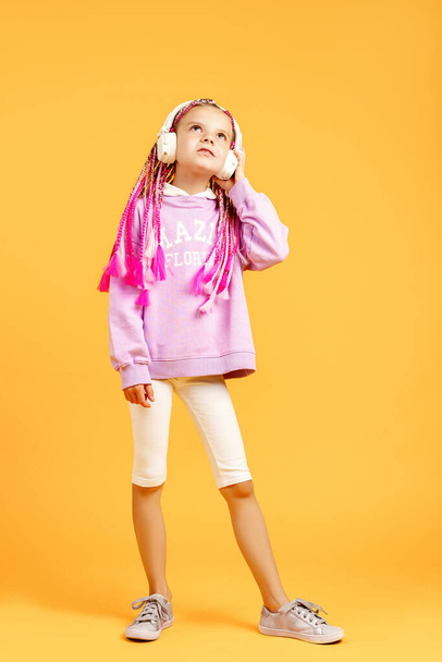 Adorable child in rounded glasses with pink dreadlocks listen to music playing in earphones and dance on yellow background. Cute small kid listening to music in headset, copy space - Photo, Image