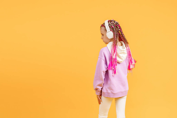 Back view of Adorable child in rounded glasses with pink dreadlocks listen to music playing in earphones on yellow background. Cute small kid listening to music in headset, copy space - Zdjęcie, obraz