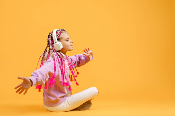 Back view of Adorable child in rounded glasses with pink dreadlocks sitting on floor and listen to music playing in earphones on yellow background. Cute kid listening to music in headset, copy space - Photo, Image