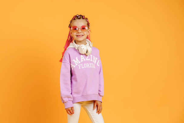 Adorable child in rounded glasses with pink dreadlocks listen to music playing in earphones on yellow background. Cute small kid listening to music in headset, copy space - Foto, imagen