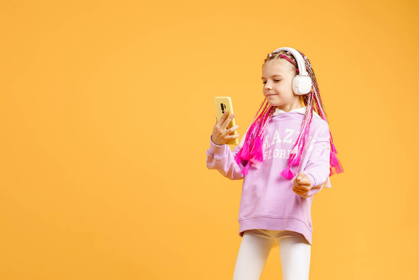 Pretty modern girl with pink dreadlocks in headphones smiling and making selfie on smartphone isolated over yellow - 写真・画像