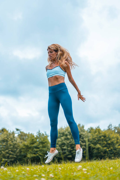 Fitness session with a young blonde caucasian woman exercising in nature with a blue maya on her feet and a white short shirt. Squatting and jumping - Foto, Imagem