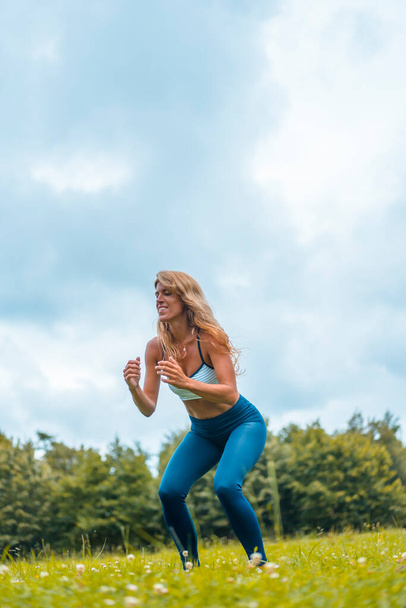 Fitness session with a young blonde caucasian woman exercising in nature with a blue maya on her feet and a white short shirt. Squatting and gathering strength to jump - Zdjęcie, obraz