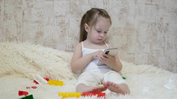 Il bambino gioca al telefono mentre è seduto sul divano con il designer. Giocattolo nello smartphone. La vita come educazione prescolare a distanza. Bambino moderno - Filmati, video