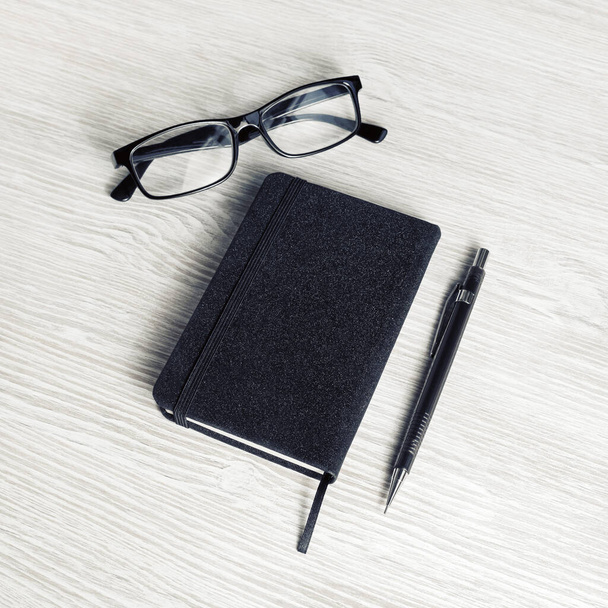 Photo of closed blank black notebook, glasses and pencil on light wood table background. - Fotografie, Obrázek