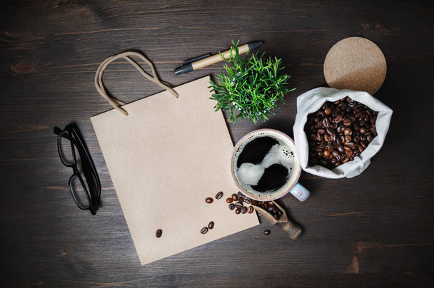 Χαρτικά και καφές Κραφτ. Vintage χάρτινη σακούλα, φλιτζάνι καφέ, κόκκους καφέ, ποτήρια, στυλό και φυτό σε ξύλινο τραπέζι φόντο. Επίπεδη. - Φωτογραφία, εικόνα