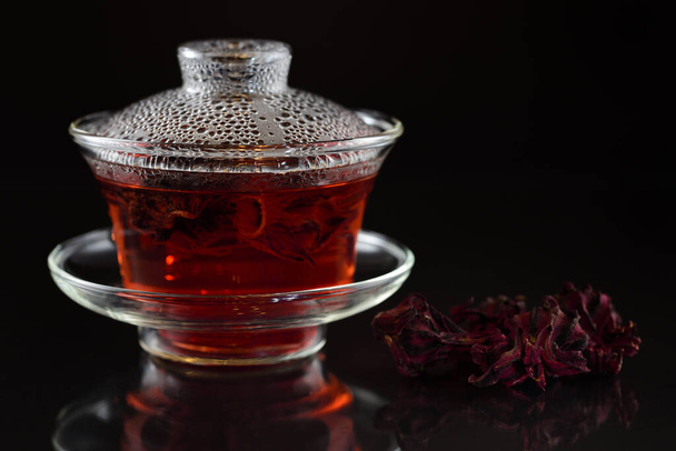 Стакан китайского чая с крышкой наполнен горячим чаем гибискуса на темном фоне и сушеные цветы рядом с ним с отражением
 - Фото, изображение
