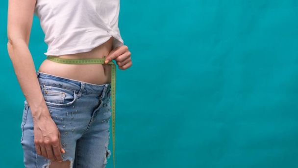 Perfectly slender slender young body of a girl. An example of sports, fitness or plastic surgery and aesthetic cosmetology. Woman measures her waist on a green background - Photo, Image