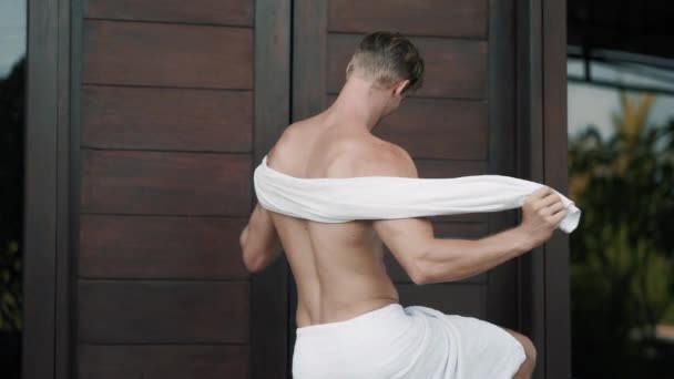 Backside view of athletic man wipes body with towel standing near wooden door - Felvétel, videó