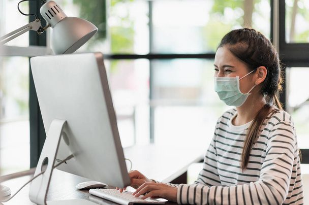 Beautiful asian woman wearing protection mask work from home in the morning. Young asian female working with computer stay at home. - Photo, Image