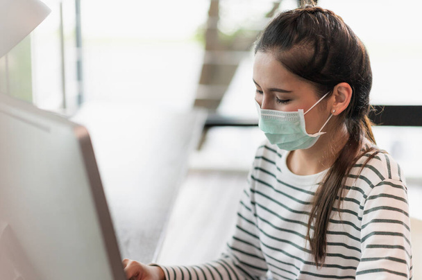 Beautiful asian woman wearing protection mask work from home in the morning. Young asian female working with computer stay at home. - 写真・画像