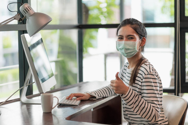 Beautiful asian woman wearing protection mask work from home in the morning. Young asian female working with computer stay at home. - Photo, Image