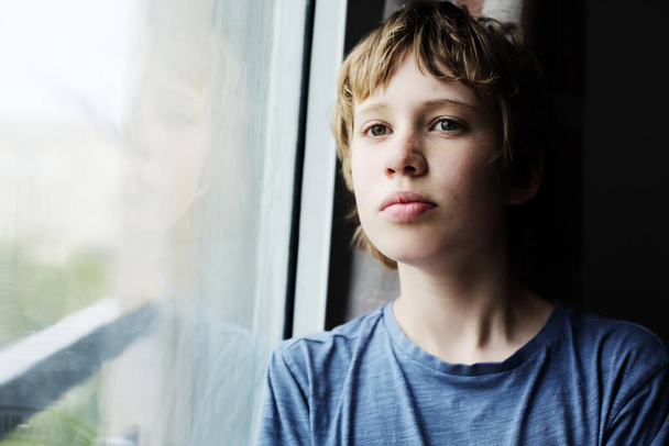 Χαριτωμένο 12χρονο αυτιστικό αγόρι κοιτάζει μέσα από το παράθυρο - Φωτογραφία, εικόνα