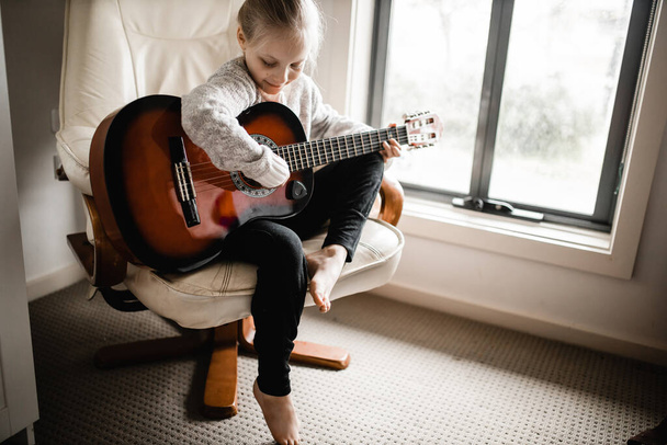 Eine junge Blondine sitzt am Fenster und übt ihre Akustikgitarre - Foto, Bild