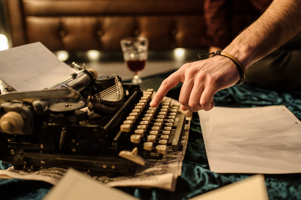 zijaanzicht van de hand van de mens typen op vintage typemachine op een stuk krant en donkerblauw fluweel stof op de vloer in een donkere kamer met gloeilampen in de buurt van een bruine leren bank - Foto, afbeelding