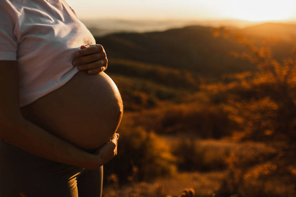 Les mains de la femme tiennent le ventre pendant la grossesse à l'extérieur gros plan. Incroyable lumière du coucher de soleil chaud. Concept de maternité et d'accouchement. - Photo, image