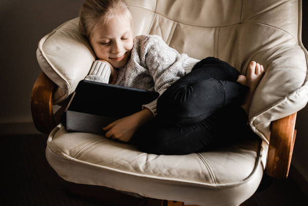 Een jong meisje van caucasion afdaling zitten door een venster kijken naar programma 's op haar tablet - Foto, afbeelding