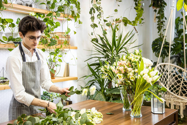 Attractive young male florist with glasses and apron works in a flower shop. pruning flower stems and preparing for bouquet assembly - Foto, Bild