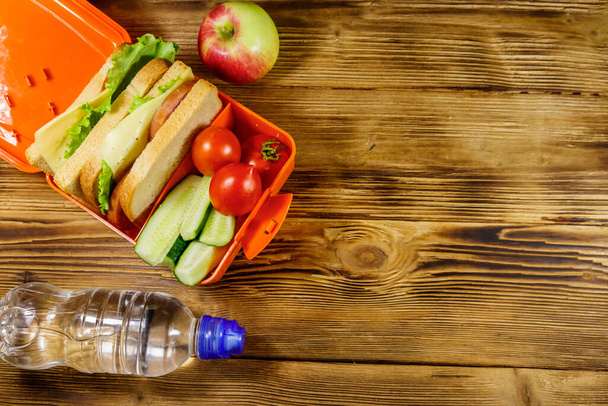 Бутылка воды, яблоко и обеденная коробка с бутербродами и свежими овощами на деревянном столе. Вид сверху, пространство для копирования - Фото, изображение