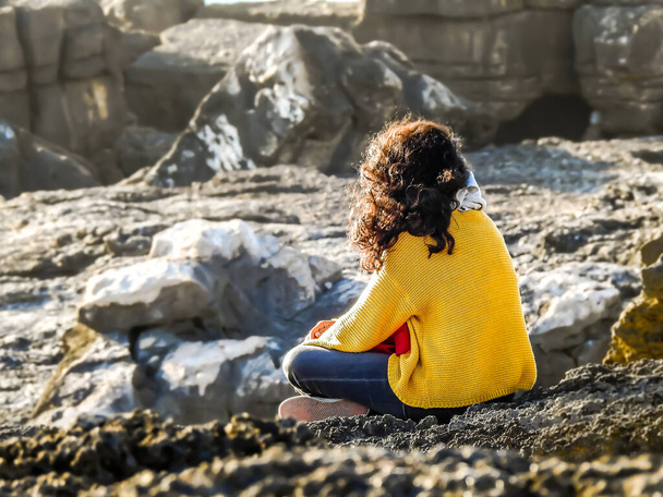 遠くを見ると海のそばの岩の上に座っている黄色のブラウスの女性 - 写真・画像