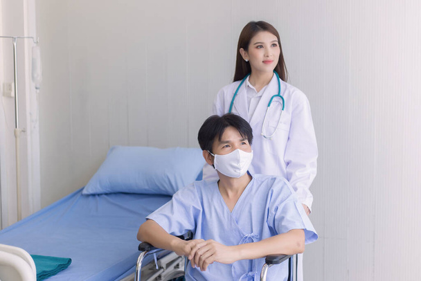 Concepto de protección contra el Coronavirus. Mujer doctora asiática hablando con un paciente hombre que usa mascarilla facial sobre su síntoma de salud mientras se sienta en silla de ruedas en el hospital. - Foto, imagen