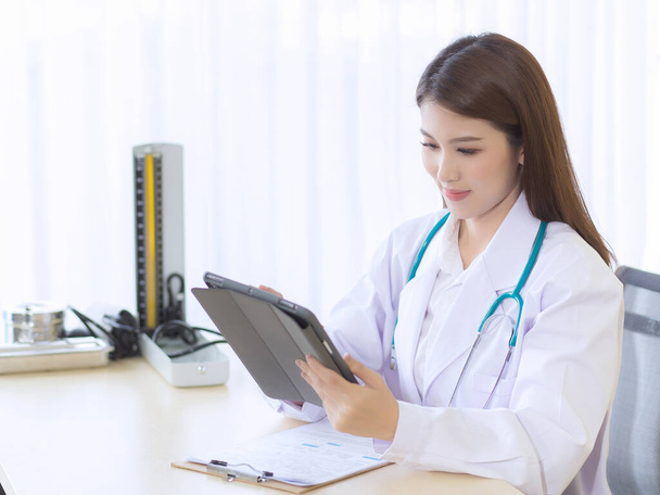 アジアの女性医師はタブレットで患者の報告書を作業している。病院のテーブルオフィスにはクリップボードと血圧モニターがあります。コロナウイルス保護の概念. - 写真・画像