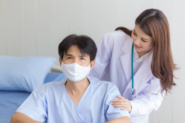 Coronavirus protection concept. Asian doctor woman talking with a man patient who wear face mask about his health symptom while he sit on wheelchair at hospital. - Photo, Image