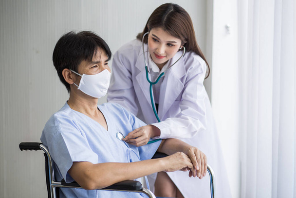 アジアの女性医師は、病院の車椅子に座りながら顔のマスクをした男性患者の心臓のリズムを確認するために聴診器を使用しています. - 写真・画像