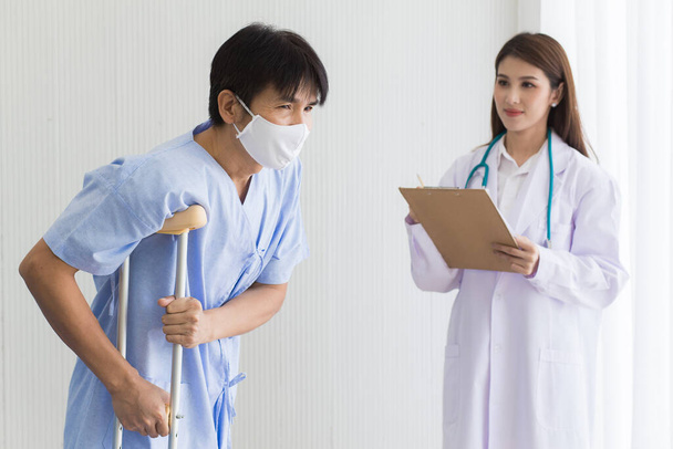 Femme asiatique médecin vérifier la marche d'un patient masculin qui porte un masque facial pendant qu'il utilise une marchette pour soutenir le stand. Elle vérifie le symptôme du patient dans un presse-papiers à l'hôpital. - Photo, image