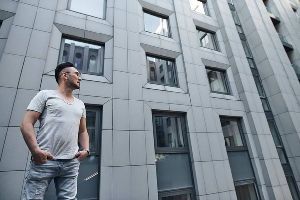 a stylish guy with glasses looks towards the office building in gray, hands in pockets. strong strict male gaze. a fashionable gray T-shirt and light jeans. portrait - Photo, Image