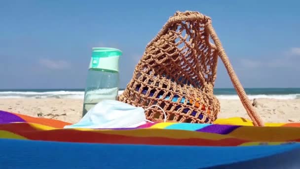 Primo piano sulla borsa da spiaggia donna, bottiglia d'acqua e maschera viso su una spiaggia di sabbia, con onde del mare sullo sfondo - Filmati, video