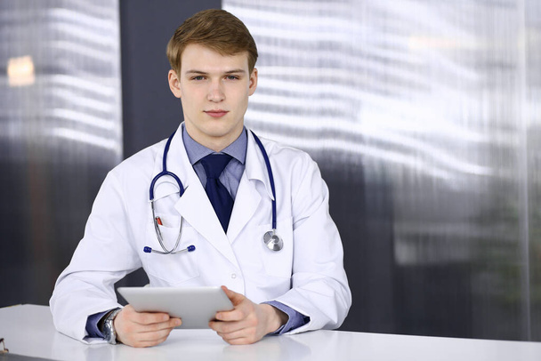 Blond male doctor sitting and working with tablet computer in clinic at his working place, close-up. Young physician at work. Perfect medical service, medicine concept - Фото, зображення