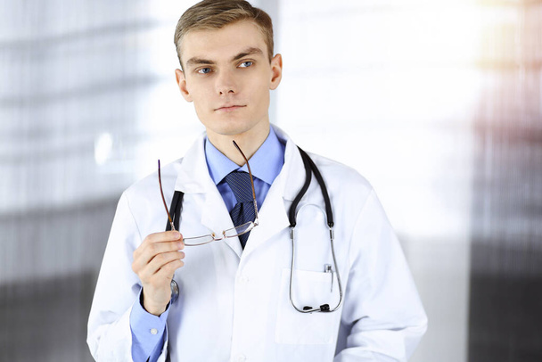 Doctor man, with glasses and stethoscope, is standing at his sunny hospital office. Perfect medical service in clinic. Happy future in medicine and healthcare - Фото, изображение