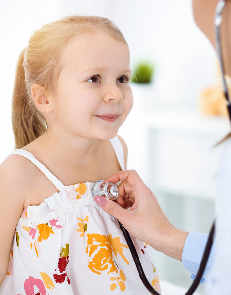 Doctor examining a child by stethoscope in sunny clinic. Happy smiling girl patient dressed in bright color dress is at usual medical inspection - Foto, imagen