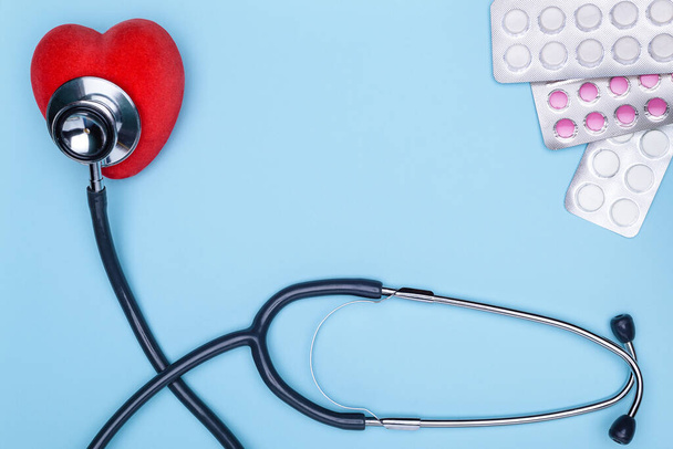 Blauer Hintergrund ist ein Statoskop, ein roter Sattel und Pillen. Das Konzept der Behandlung von Herz-Kreislauf-Erkrankungen. Nahaufnahme von bemaltem Herz, vielen Drogen und Stethoskop auf blauem Hintergrund. - Foto, Bild