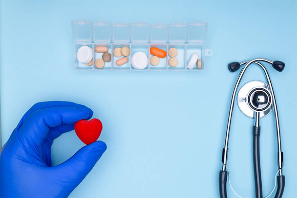 Doctor cardiologist hands with heart in hands on blue background.Pill box with medicines on blue background. Concept of cardiology and cardiovascular diseases.Statoscope on blue background. - Фото, изображение