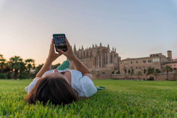 jeune fille influenceuse couchée sur l'herbe sur le dos en utilisant son téléphone portable profitant d'un coucher de soleil doré devant la cathédrale de Palma de Mallorca, fille brune aux cheveux bruns et au ciel clair - Photo, image